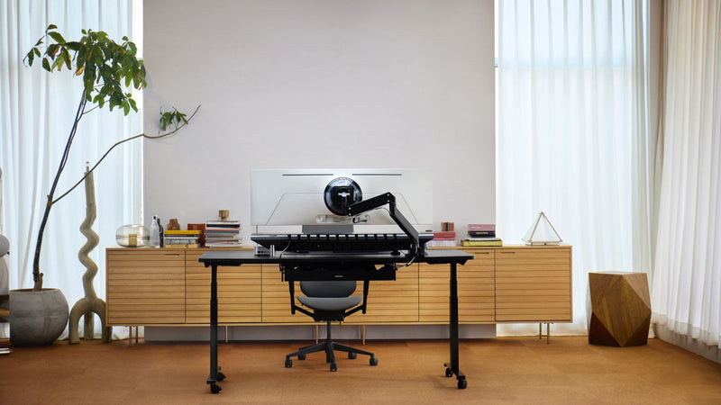 A minimalist office setup with a black standing desk, ergonomic chair, and a monitor on an adjustable arm. A wooden storage unit, plant, and decorative items complete the clean, modern look.