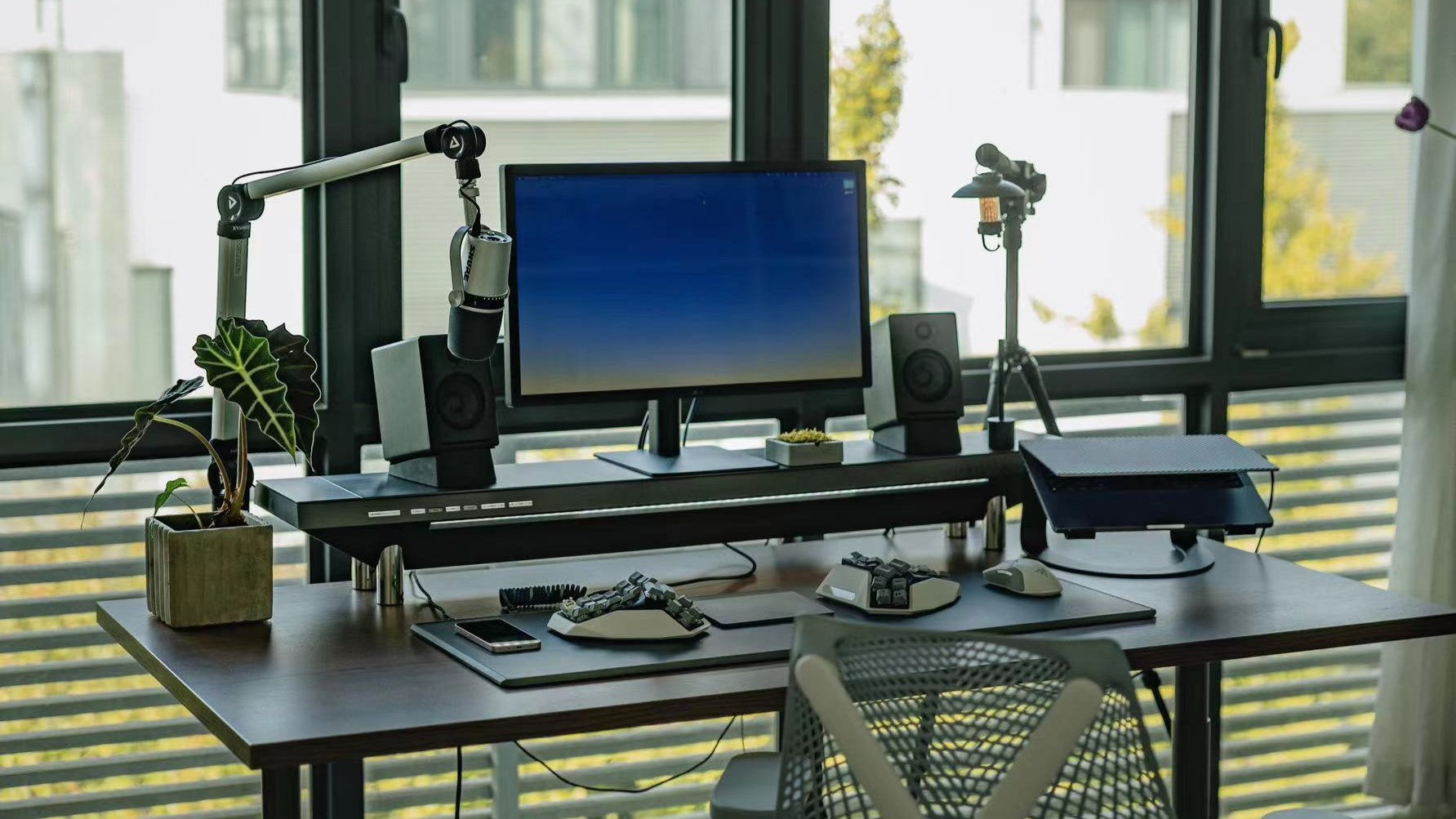 An advanced ergonomic workstation setup featuring the Hexcal Studio, equipped with a monitor, speakers, microphone arm, and additional tech accessories, all organized on a sleek desk with a modern aesthetic and natural lighting in the background.
