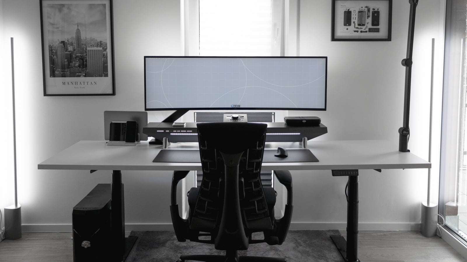 A minimalist and modern ergonomic workstation featuring an ultrawide monitor mounted on the Hexcal Studio, accompanied by sleek tech accessories, a comfortable ergonomic chair, and a clean, organized desk space. The setup is enhanced by ambient lighting and monochrome wall art in a bright, well-lit room.